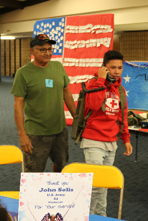 Military veteran looks on as a junior high boy uses a phone attached to a backpack Military veteran looks on as a junior high boy uses a phone attached to a backpack